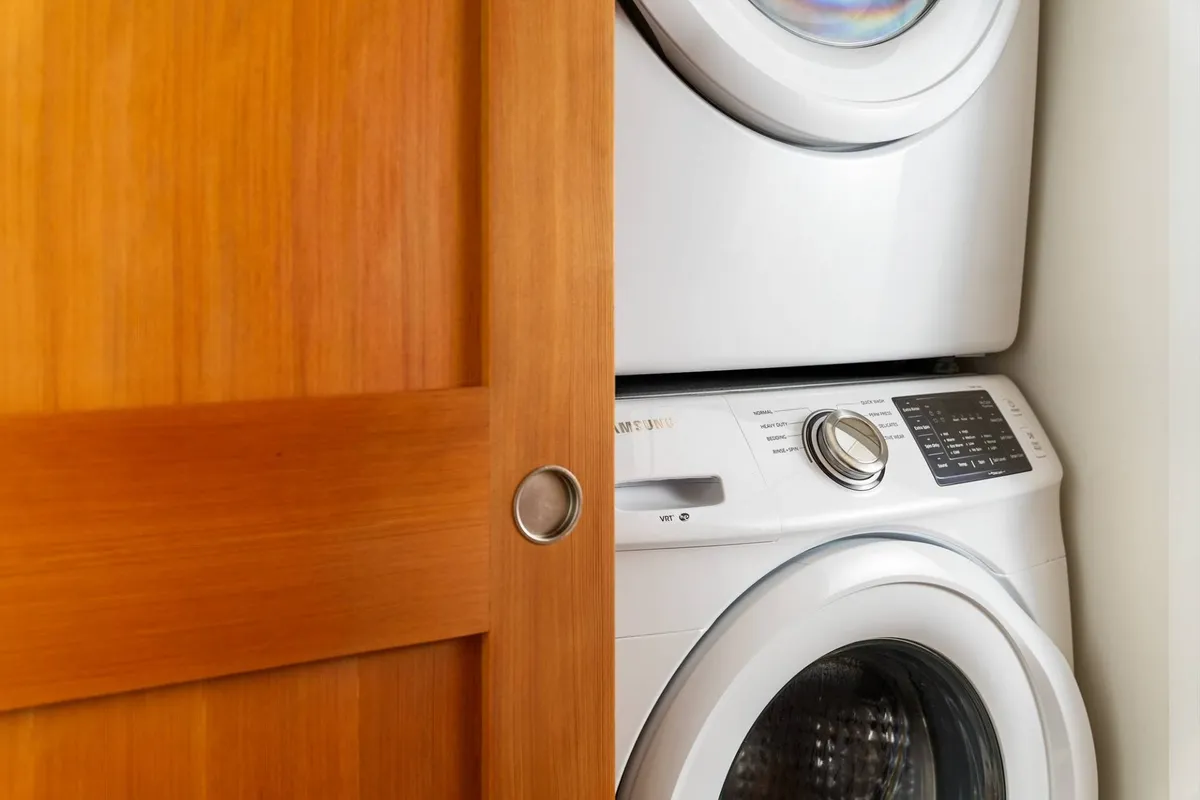 A well-lit laundry area with a stacked washer and dryer sits neatly beside a warm wooden door. The space is uncluttered and efficient, offering all the essentials for handling laundry with ease.