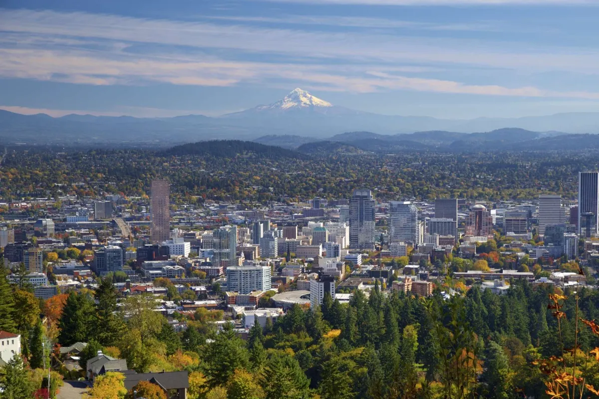 A panoramic view of Portland’s downtown skyline framed by lush green trees, with a snow-capped mountain rising majestically in the background. The image evokes a sense of proximity to both urban excitement and natural beauty, showcasing the city as vibrant and accessible.