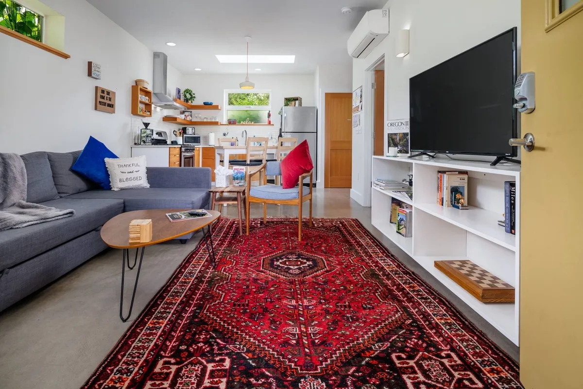 This image displays an inviting living room area with a modern sofa, a stylish rug, a view toward the dining area, and a mounted television. The natural light pouring in accentuates the clean, contemporary lines of the furnishings and the open-concept layout.