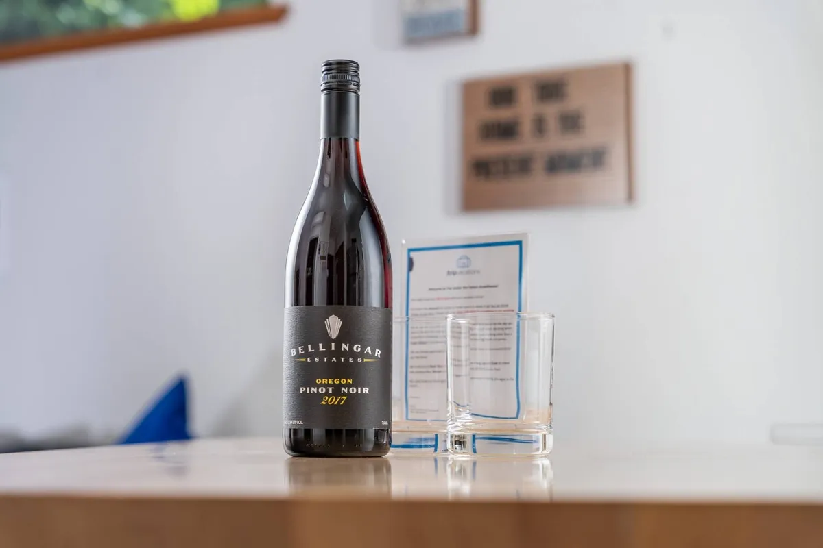 A welcoming setup with a bottle of Bellingar Estates Pinot Noir, two empty glasses, and an informational pamphlet, suggesting the opportunity for socializing, relaxing, or learning about local offerings in a comfortable, communal space.