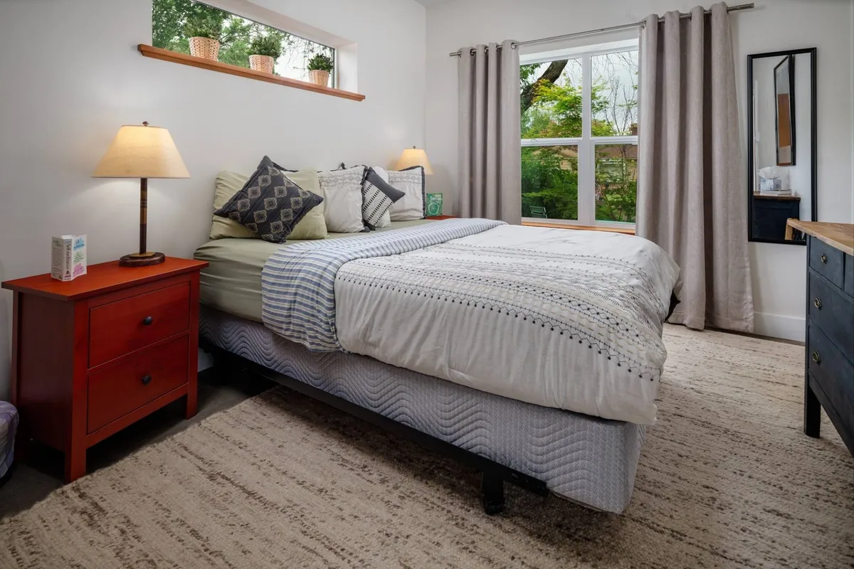 A tranquil bedroom scene featuring a neatly made queen-size bed with plush bedding and decorative pillows. Soft natural light pours in from a large window adorned with elegant curtains, and a cozy nightstand adds warmth to the inviting space.