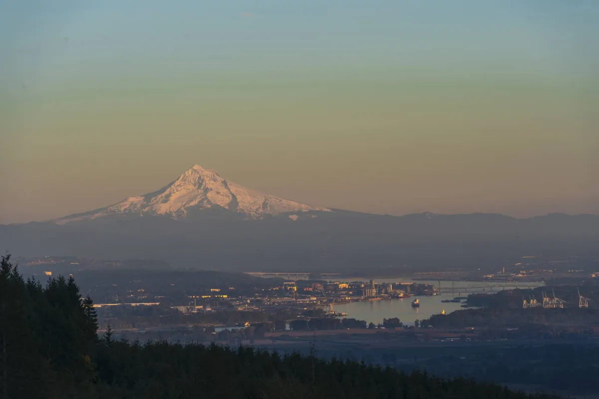 This image captures a tranquil view of Mount Hood in the distance, with a river in the foreground, surrounded by forest and hints of an industrial area. The landscape is bathed in natural light, evoking feelings of serenity and connection to the outdoors.