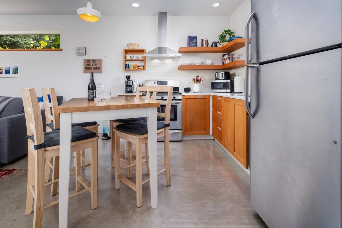 A bright dining area, seamlessly connected to the kitchen, features a modern dining table, sleek kitchen appliances, and shelves adorned with kitchenware. The room feels casual and welcoming, ideal for shared meals or an evening in.
