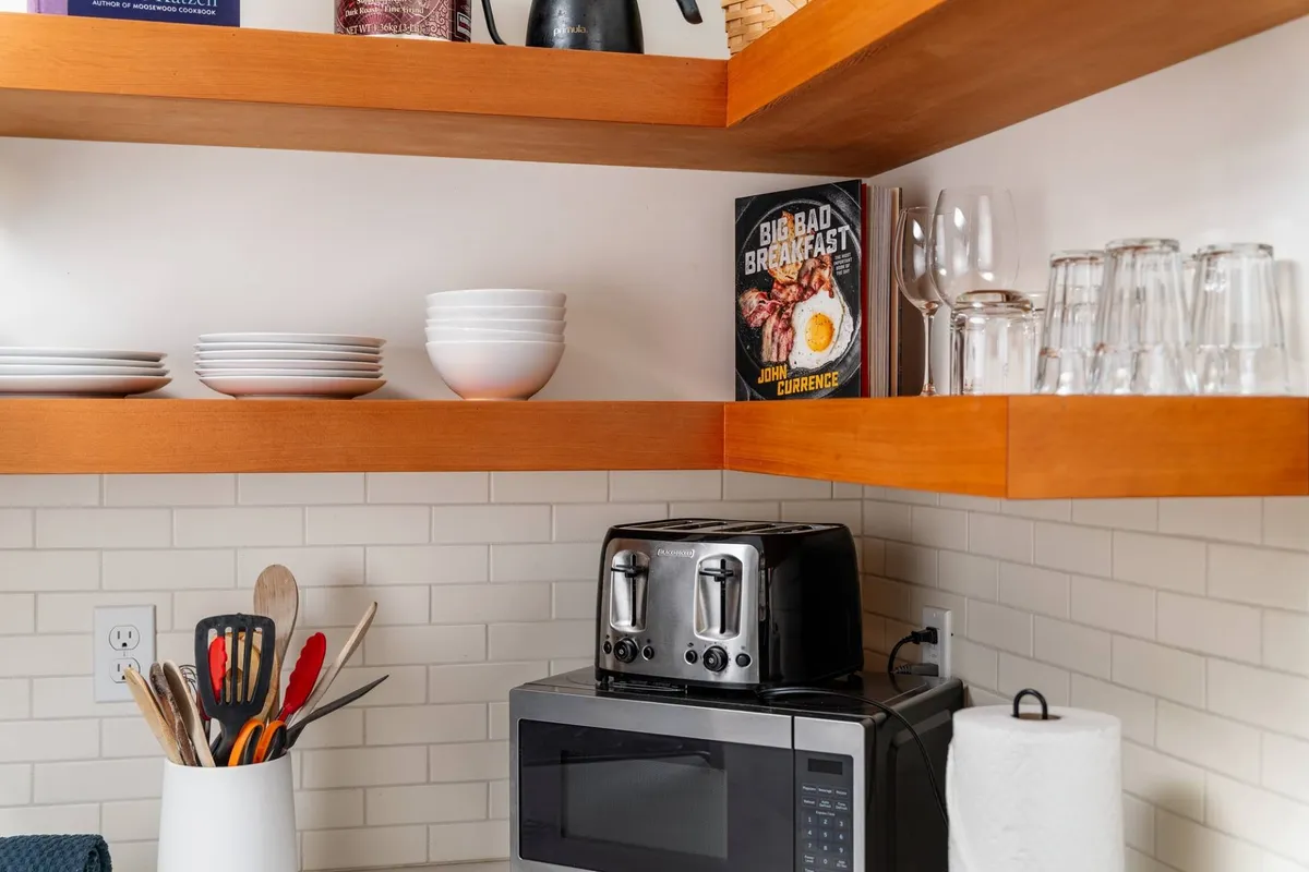 A homey kitchen view with a toaster, microwave, cookware, stacked plates, and an open cookbook, all arranged on and around generous counter space. The scene suggests active cooking and meal preparation, evoking the idea of entertaining and experimenting with new recipes.